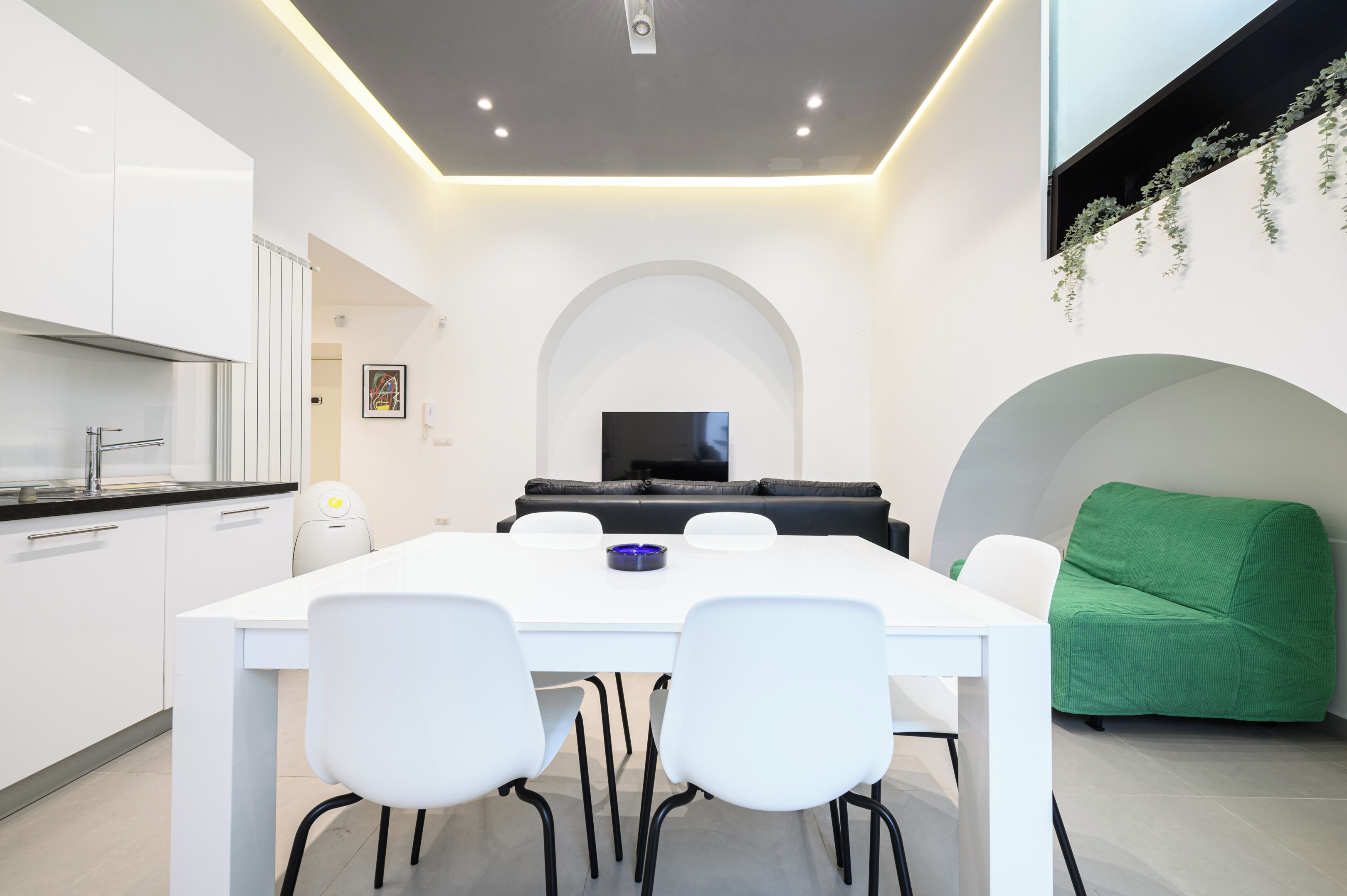 Una cucina moderna con mobili bianchi e un piano di lavoro nero, una zona pranzo con tavolo e sedie bianche, un divano nero e una poltrona verde.