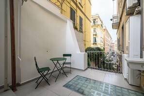 A balcony with a glass table and two chairs, a white wall, and a view of a narrow street with buildings. - Via Morghen 63 (Naples)