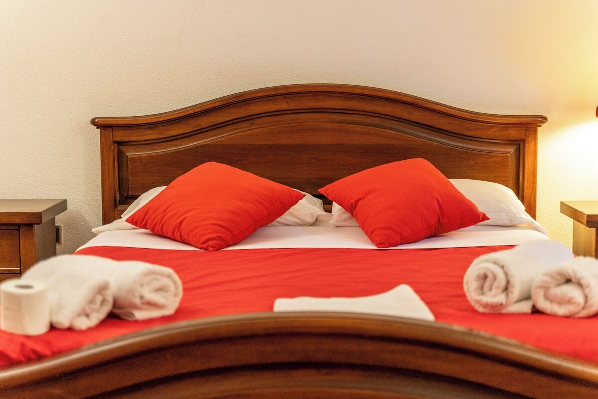 A neatly made bed with red bedding and two red pillows, a wooden headboard, and a nightstand with a lamp.