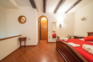Quarto com cama de madeira, uma pequena mesa de cabeceira, um espelho e porta que dá para outro cômodo.