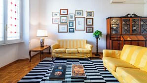 Een woonkamer met een geruit tapijt, twee gele banken, een houten kast en ingelijste foto's aan de muur.