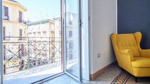 Ein Zimmer mit einem gelben Sessel, einer großen Schiebetür zum Balkon und Blick auf benachbarte Gebäude.