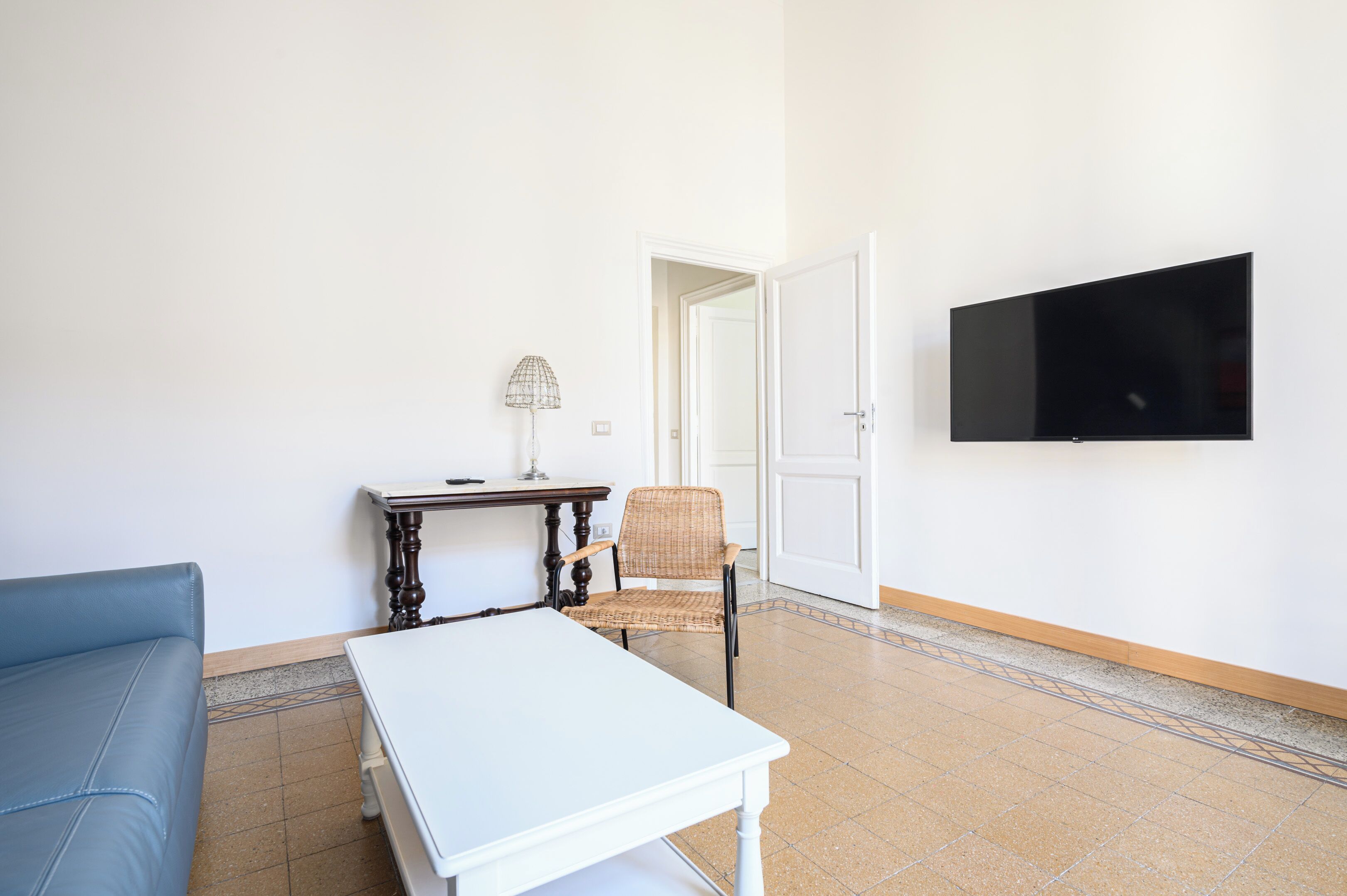 A living room with a white sofa, a small table with a lamp, a wicker chair, and a flat-screen TV mounted on the wall.