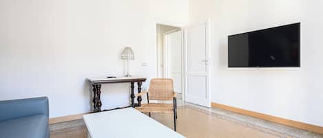 Un salon avec un canapé blanc, une petite table avec une lampe, un fauteuil en osier et un téléviseur à écran plat fixé au mur.
