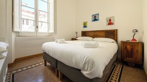 Ein ordentlich bezogenes Bett mit weißen Leinentüchern und gerollten Handtüchern, ein Holz-Kopfteil, ein Nachttisch mit Lampe und ein Fenster mit Blick auf Gebäude.