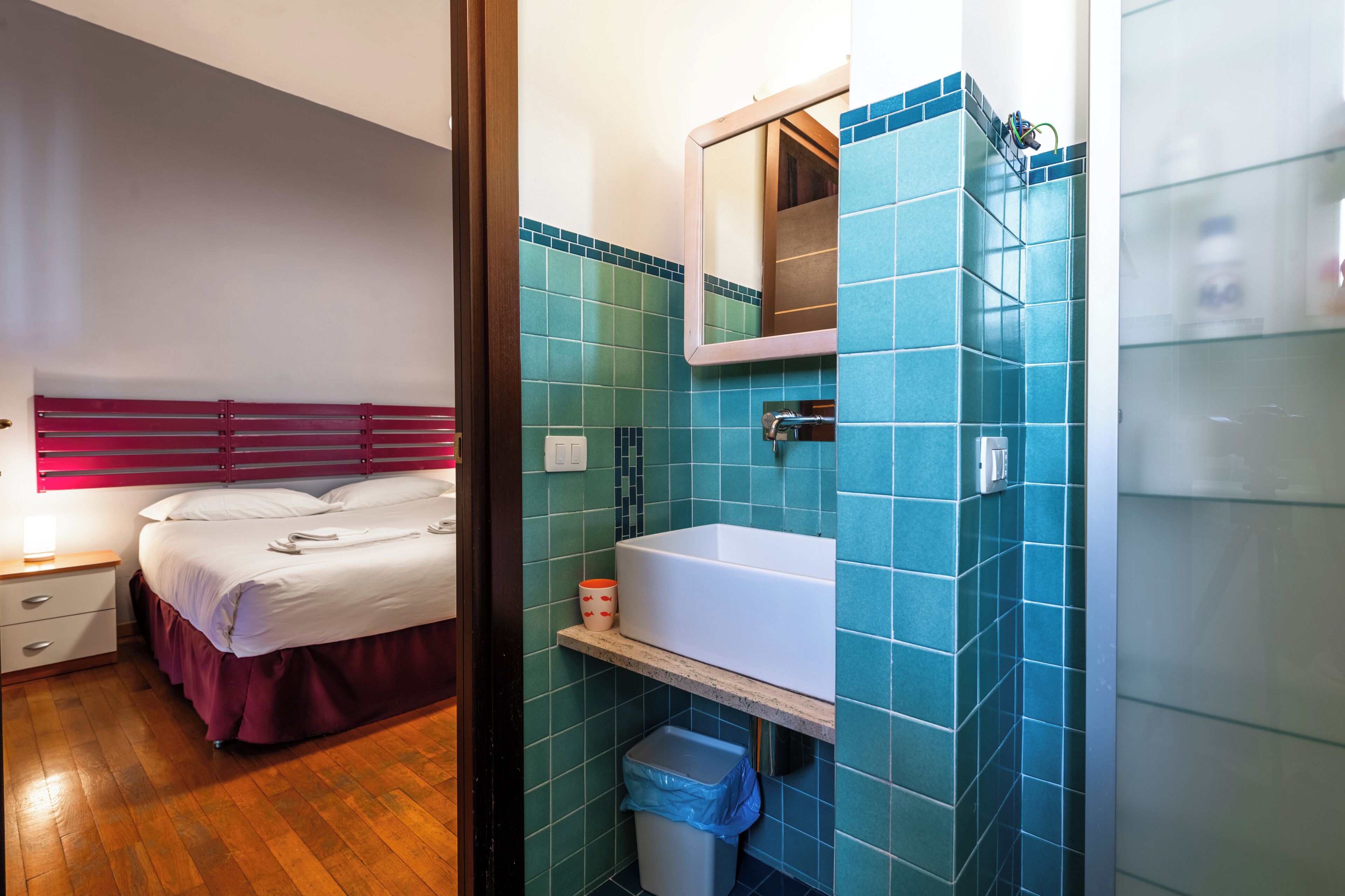 Une chambre d’hôtel avec un lit, une table de chevet, une salle de bain avec un lavabo et un miroir, et des murs recouverts de carreaux bleus.