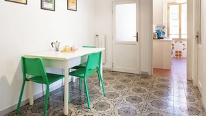 Un'area pranzo con un tavolo bianco e due sedie verdi, una porta bianca, piastrelle a pavimento decorate e quadri appesi al muro.