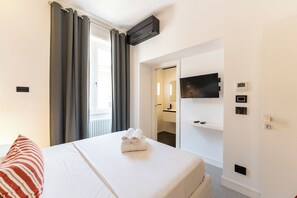 Ett hotellrum med en säng, en tv som är monterad på väggen, en dörr som leder till ett badrum och ett fönster med gardiner.