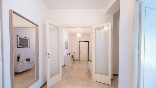 A hallway with white walls, a tiled floor, and doors leading to other rooms. There are framed pictures on the walls and a small plant on a shelf. - Casa Tripolitania (Rome)