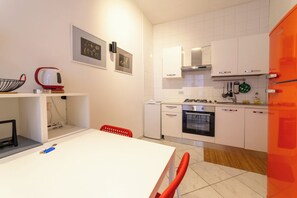 A modern kitchen with white cabinets, a red kettle, and a dining area with a white table and red chairs. - Vico Piazza Nuova 1 (Naples)