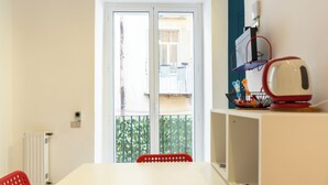 Um quarto com uma mesa branca, uma cadeira vermelha, um radiador, uma janela com vista para uma varanda e uma estante com um rádio vermelho e branco.