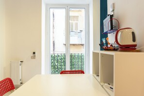 A room with a white table, a red chair, a radiator, a window with a view of a balcony, and a shelf with a red and white radio. - Vico Piazza Nuova 1 (Naples)
