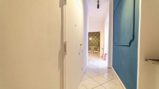 Um corredor com paredes brancas, uma porta azul e um chão de azulejos que leva a uma sala de jantar com toalha de mesa estampada.