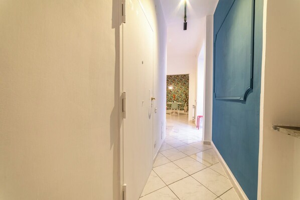 A hallway with white walls, a blue door, and a tiled floor leading to a dining area with a patterned tablecloth. - Vico Piazza Nuova 1 (Naples)