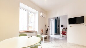 Um quarto moderno e bem iluminado, com uma mesa redonda e cadeiras, uma TV fixada na parede, e vista para a cozinha através de uma porta aberta.