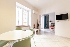 A modern, well-lit room with a round table and chairs, a TV mounted on the wall, and a view of the kitchen area through an open door. - Vico Piazza Nuova 1 (Naples)