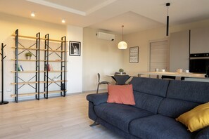 A modern living room with a grey sofa, a wooden bookshelf, a dining area, and a kitchen. - Cinecittà Dreams (Rome)