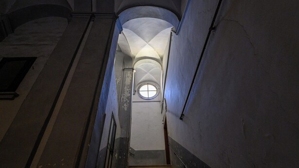 Un escalier étroit, surmonté d’une fenêtre circulaire, menant à un étage supérieur.