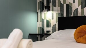 Un lit bien fait, avec un oreiller blanc et une serviette blanche roulée posée sur le côté. On aperçoit une table de chevet noire avec une lampe. Le mur est orné d’un motif géométrique.