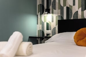 Una cama bien hecha con una almohada blanca y una toalla blanca enrollada al lado. Se ve una mesita de noche negra con una lámpara. La pared tiene un diseño geométrico.