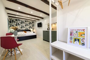 A modern bedroom with a bed, bedside table, and a wall-mounted TV. There is a dining area with a table and chairs, and a kitchen area with cabinets and a countertop. - La Stanza del Principe (Naples)