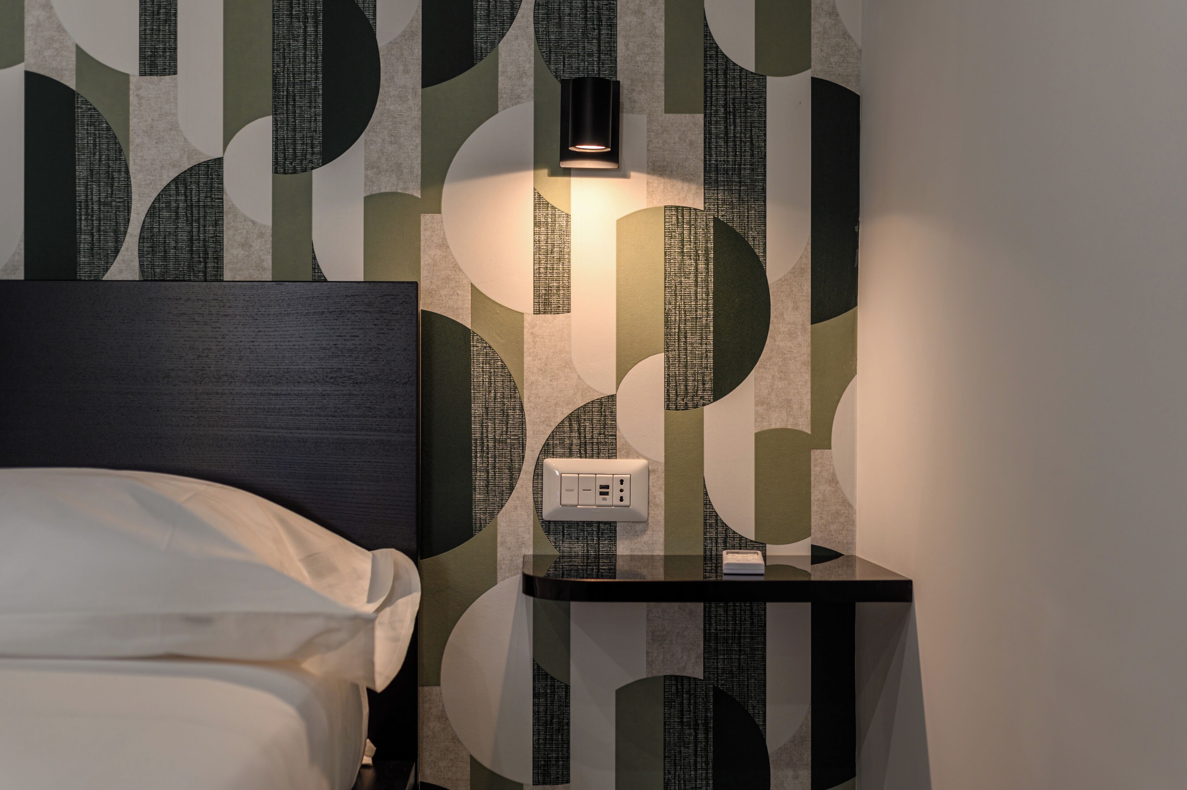 Ein ordentlich bezogenes Bett mit dunkelbrauner Kopfteile, ein Nachttisch mit Lampe und einer Steckdose. Die Wand ist mit einem geometrischen Muster tapeziert.