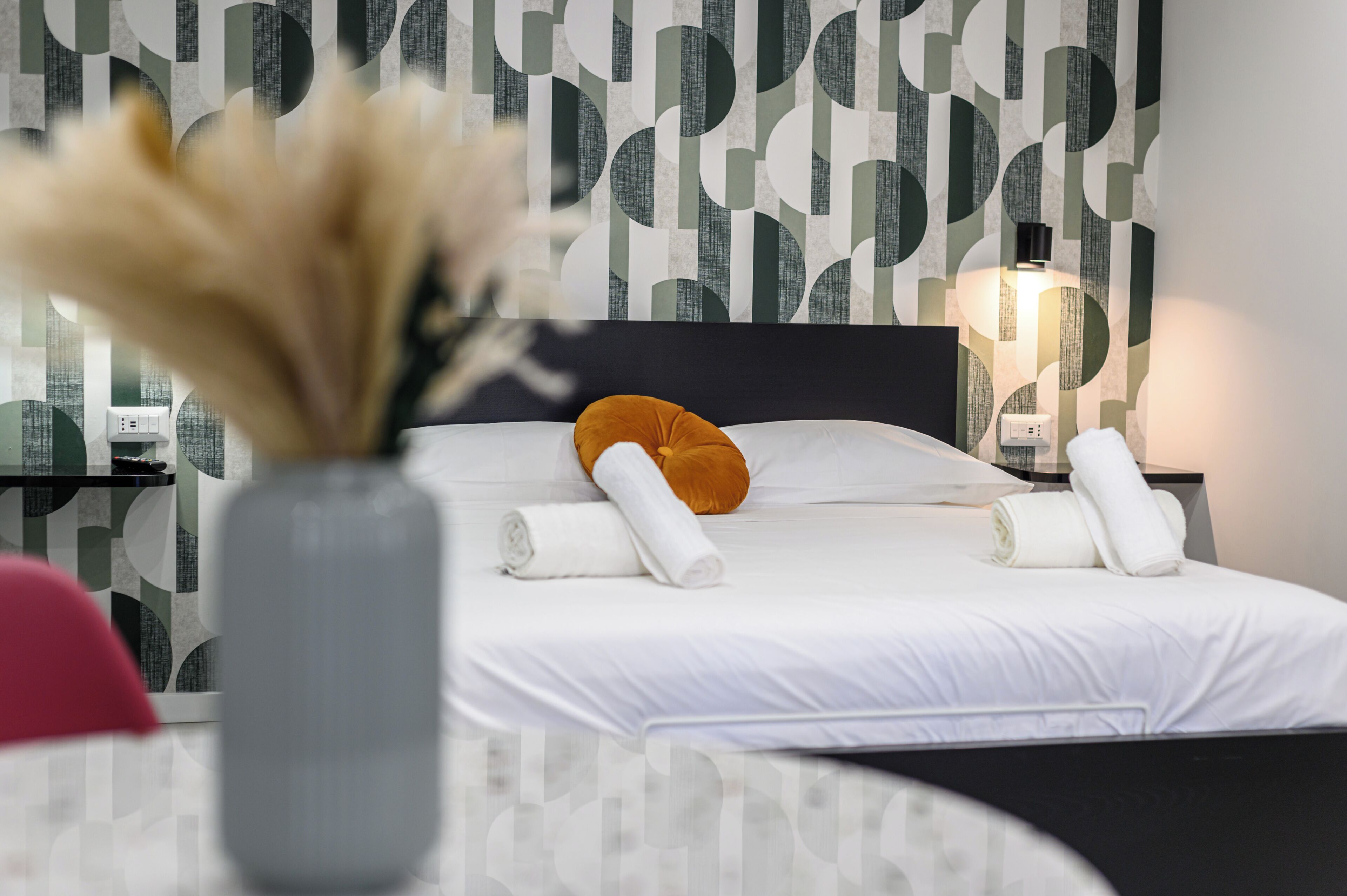 Een net opgemaakt bed met witte lakens en een oranje sierkussen, een vaas met gedroogde bloemen, en een muur met een geometrisch patroon.