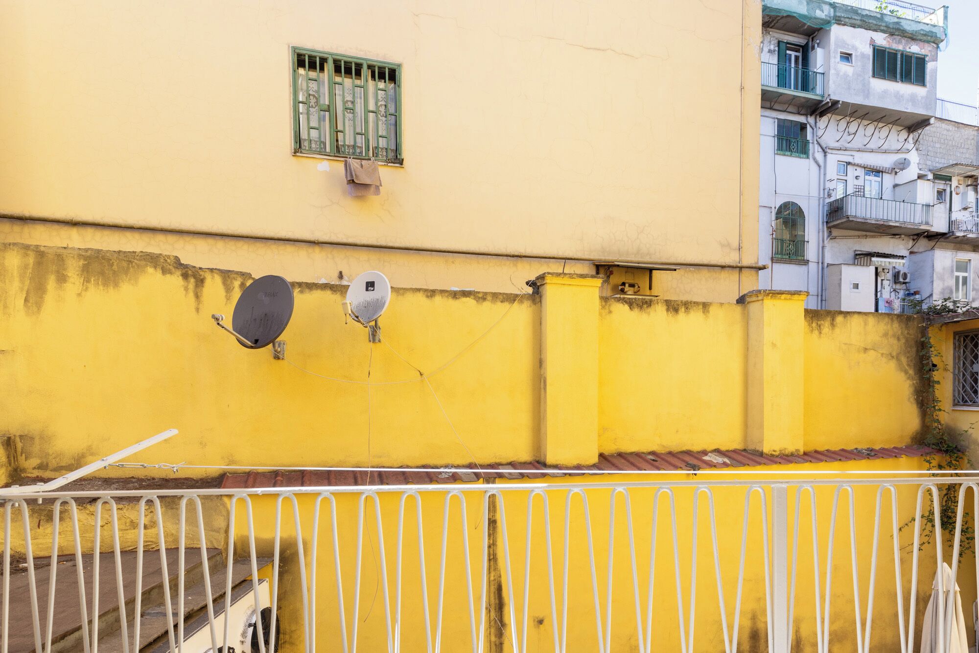 A yellow building with satellite dishes, a window with green bars, and a metal fence.