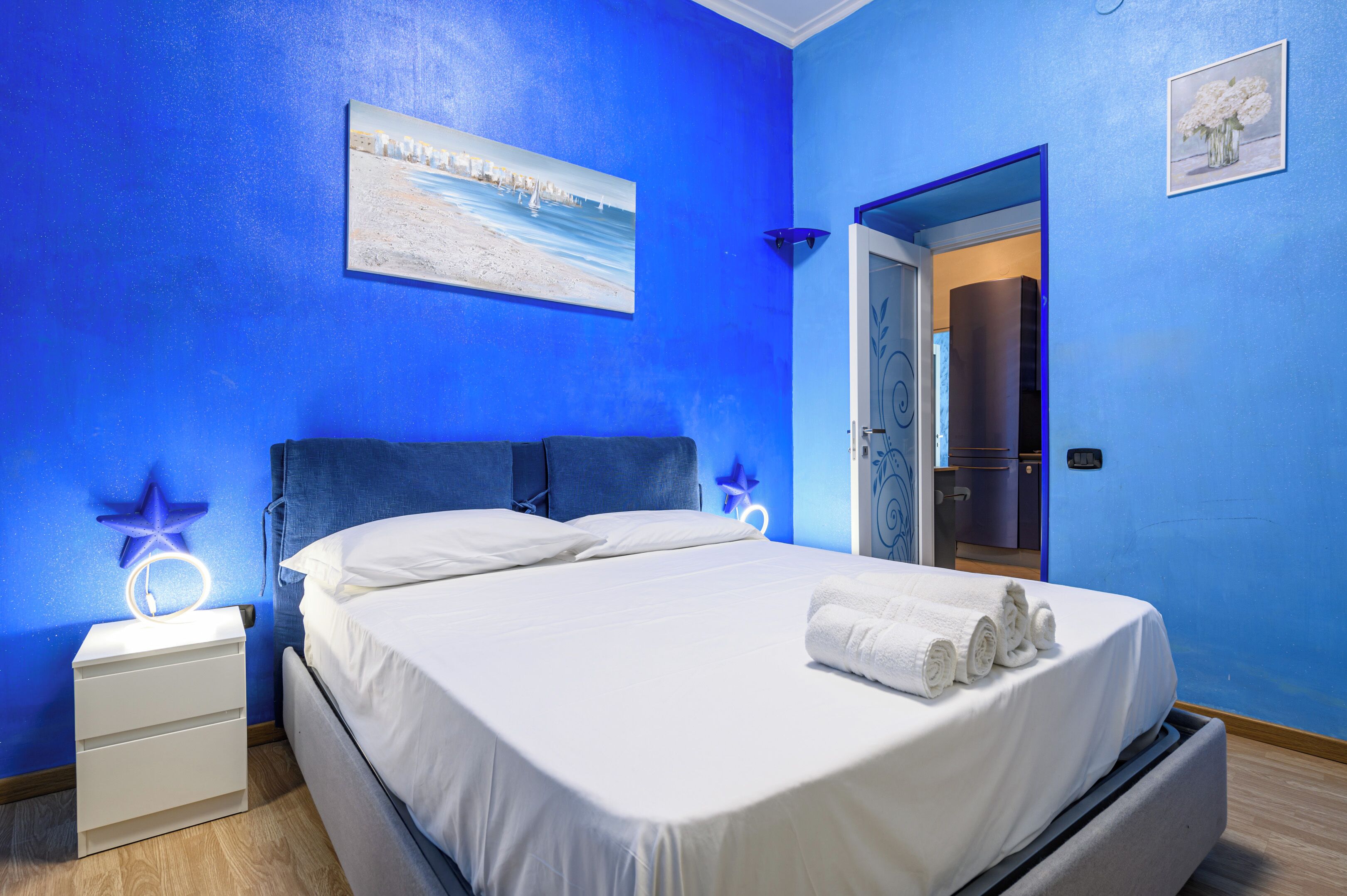 Une chambre à coucher avec un lit, du linge de lit blanc, une table de chevet avec une lampe et un tableau au mur.