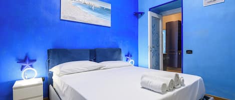 A bedroom with a bed, white bedding, a nightstand with a lamp, and a painting on the wall.