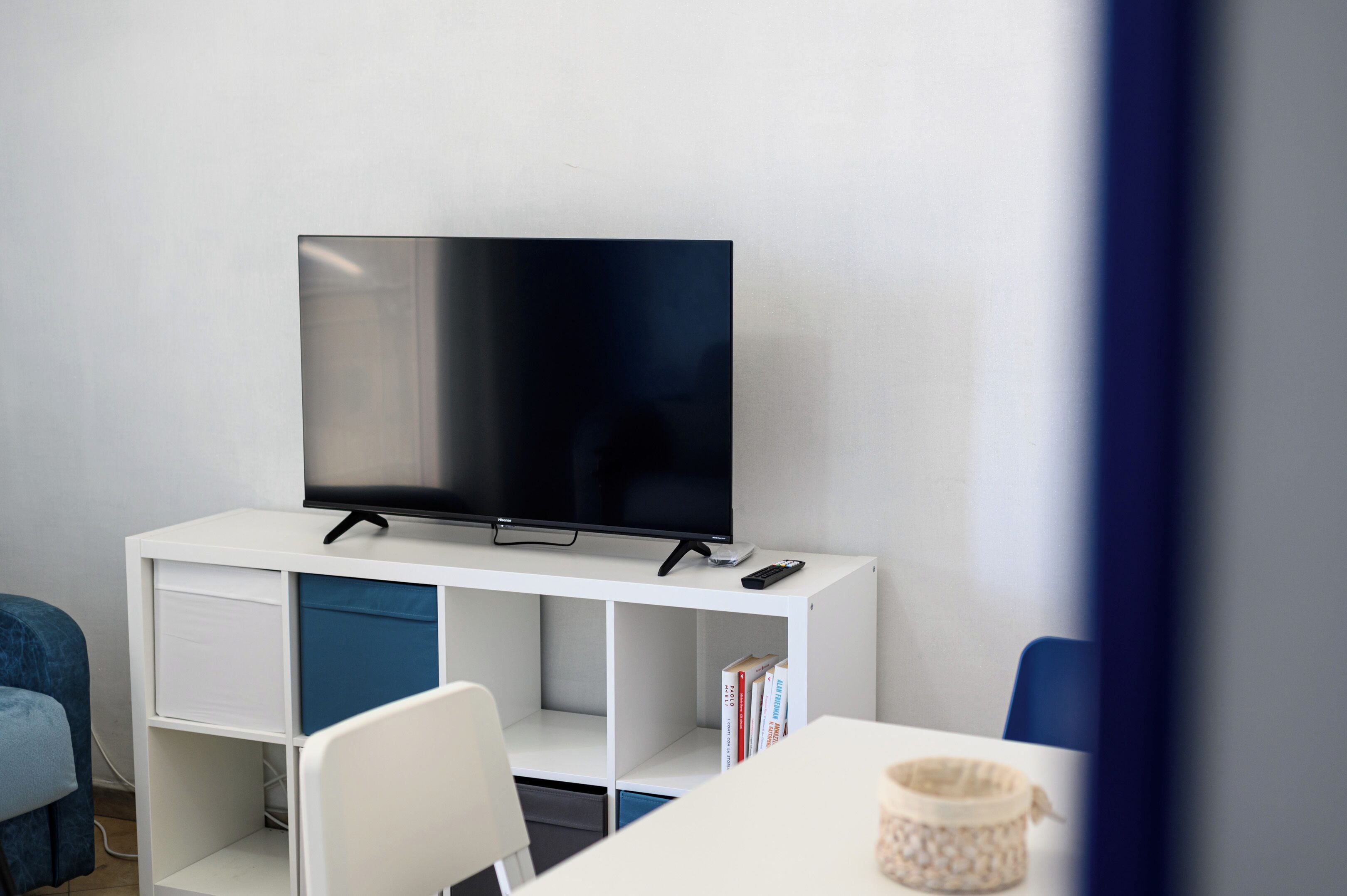 Una camera moderna con una TV a schermo piatto su un mobile bianco, una scrivania bianca con una sedia e una poltrona blu.