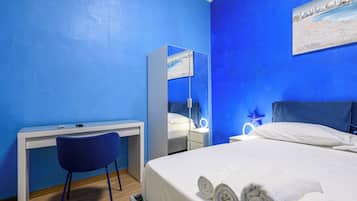 Una camera d'albergo con un letto, una scrivania, una sedia, uno specchio e un comodino con una lampada.