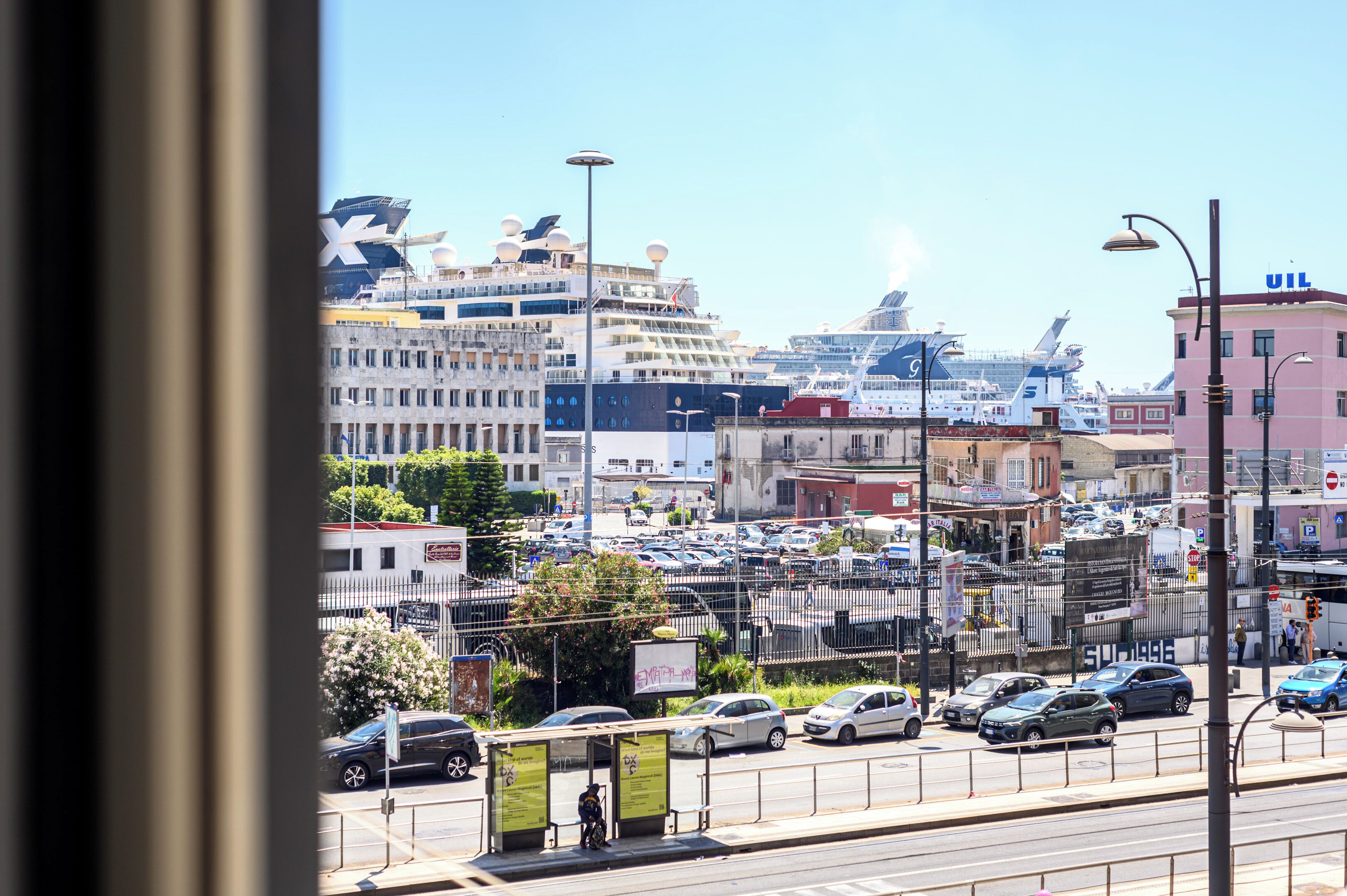 A busy urban street with parked cars, a bus stop, and a large cruise ship docked in the background.
