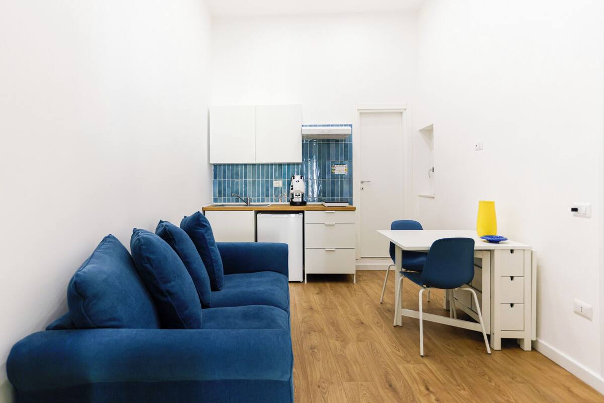 Ein kompakter Wohnraum mit einem blauen Sofa, einer kleinen Küchenzeile, einem weißen Schreibtisch und einem gelben Blumenvasen.