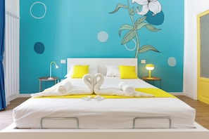 Habitación de hotel con cama, dos mesitas de noche, una lámpara de noche, un mural en la pared y una puerta.