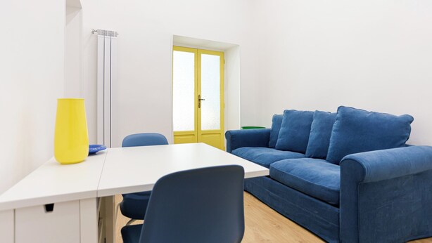 Een lichte, kleine kamer met een wit bureau, een blauwe bank en een gele vaas.