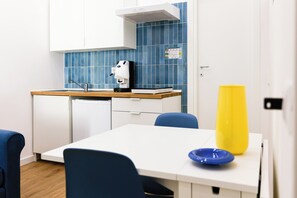 Een moderne keuken met witte kasten, een blauwe betegelde spatwand, een koffieapparaat en een gele vaas op het aanrecht.