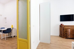 Een kamer met een gele deur, een witte muur, een houten dressoir, een aan de muur bevestigde televisie en een bureau met stoel.