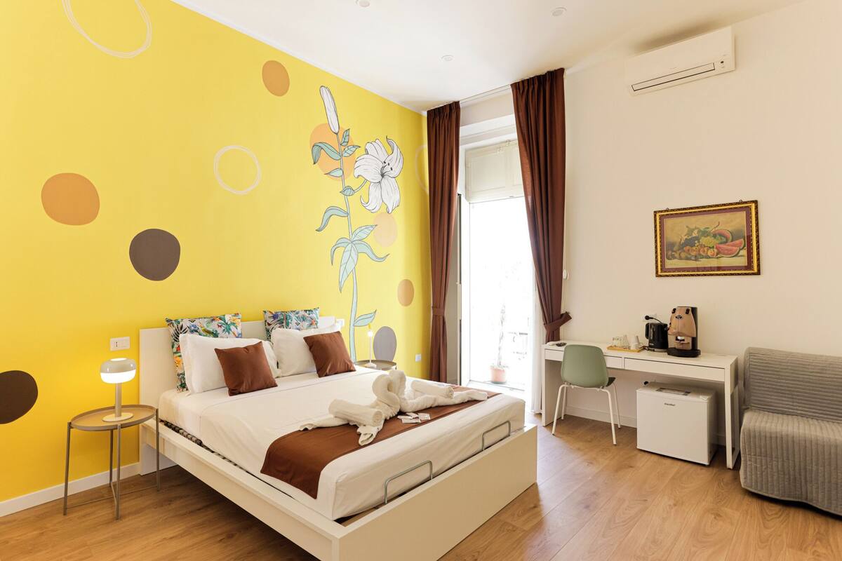 Ein Hotelzimmer mit Bett, Schreibtisch und Stuhl. Ein Gemälde hängt an der Wand und ein Klimagerät ist vorhanden.