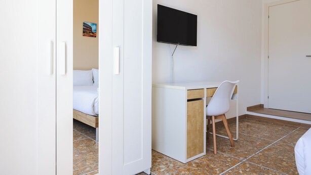 Un dormitorio con una cama, un escritorio con una silla, un televisor montado en la pared y una puerta.