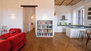 A modern living room with a red sofa, a bookshelf, and a kitchen area with a wooden herringbone floor. - Boho 202 (Rome)