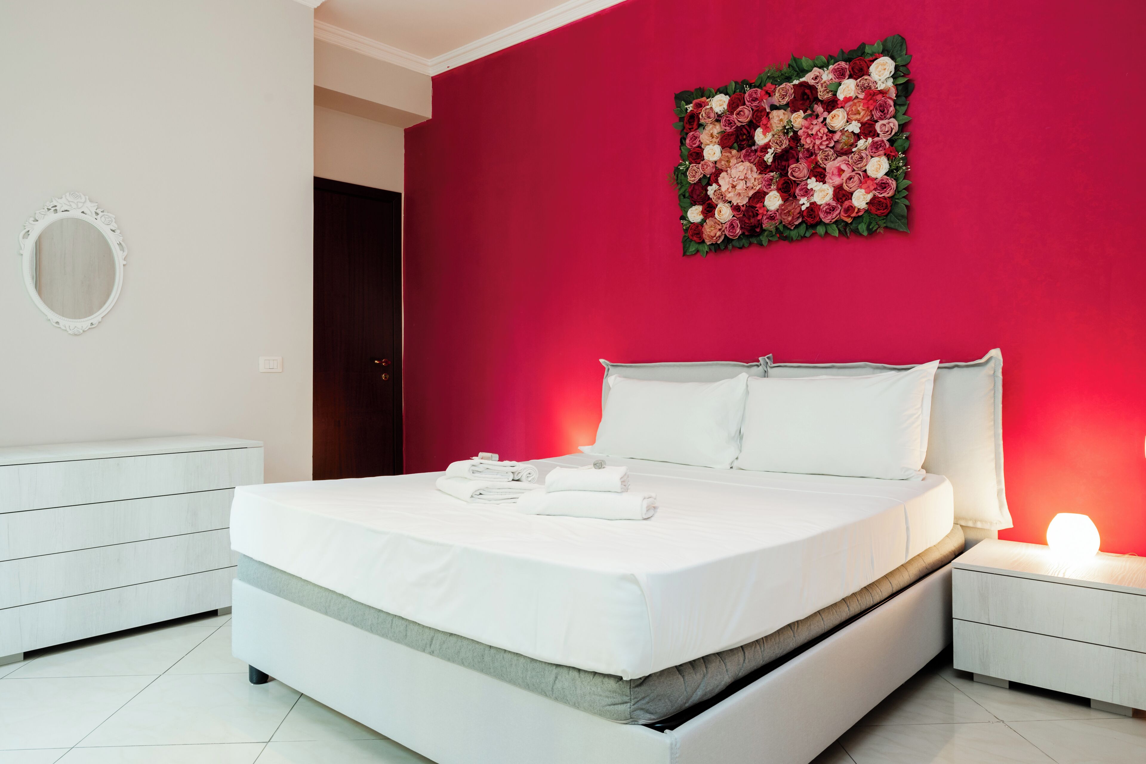 Ein modernes Schlafzimmer mit einem großen Bett, weißer Bettwäsche, einem Nachttisch mit Lampe, einer Kommode und einem floralen Wandbild.