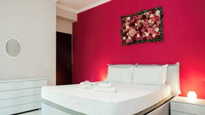 Ein modernes Schlafzimmer mit einem großen Bett, weißer Bettwäsche, einem Nachttisch mit Lampe, einer Kommode und einem floralen Wandbild.