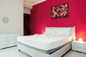 A modern bedroom with a large bed, white bedding, a nightstand with a lamp, a dresser, and a floral wall art piece. - Romantic Malatesta (Rome)