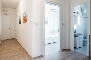 Un pasillo con piso de madera, una puerta blanca y un baño a la vista a través de una puerta abierta.