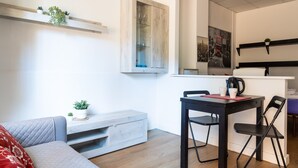 Ein modernes Wohnzimmer mit grauem Sofa, einem Holztisch und einem Essbereich mit schwarzem Tisch und Stühlen. Auf dem Tisch steht eine kleine Pflanze und darüber auf dem Regal noch eine weitere.