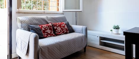 Una sala de estar moderna con un sofá gris, una ventana con vistas a vegetación, una cortina blanca y un estante de madera con plantas.