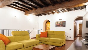 Un salon avec deux canapés, une table basse et un téléviseur. Le plafond est orné de poutres en bois et un tableau est accroché au mur.