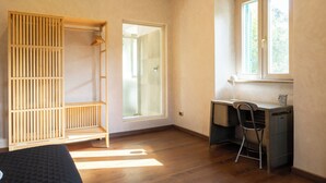A room with a wooden wardrobe, a desk with a chair, a window, and a door leading to a shower area.