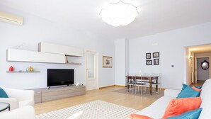 Ett modernt vardagsrum med en platt-TV, en matplats med ett träbord och stolar samt en vit soffa med färgglada kuddar.